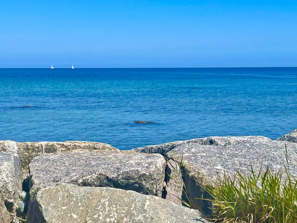 Zwei Segelboote in der Ferne auf ruhigem Meer, gesehen von der ruhigen Küste aus. Ein perfekter Junitag.